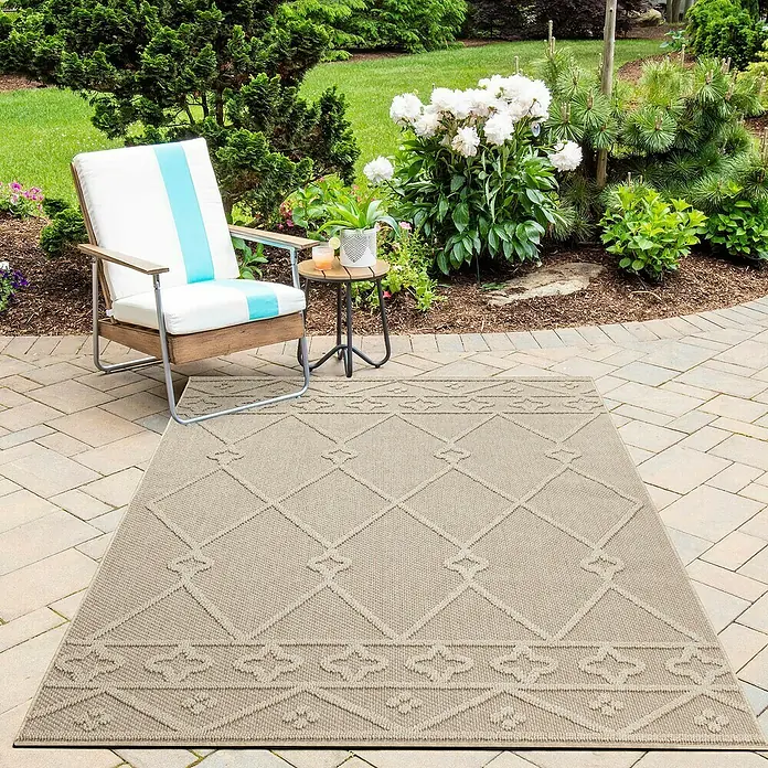 Outdoor-Teppich Patara II - Beige, 150 x 80 cm, 100 % Polypropylen - Ein beiger Outdoor-Teppich mit einem geometrischen Rautenmuster und floralen Elementen liegt auf einer Steinterrasse neben einem Sessel und einem Beistelltisch.
