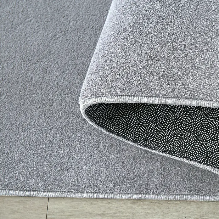 Kurzflorteppich SkySilber, 340 x 240 cm, 100 % Polyester - Detailaufnahme eines grauen Teppichs mit gewellter Ecke, die die rutschfeste Unterseite zeigt.