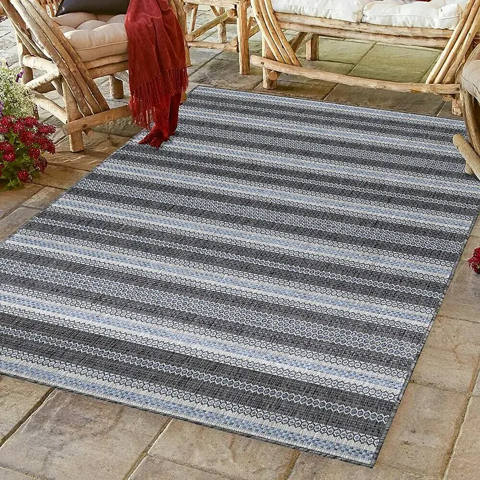 Outdoor-Teppich Sunny V - Schwarz, 170 x 120 cm, 100 % Polypropylen - Ein rechteckiger Teppich mit einem Streifenmuster aus Grau- und Blautönen liegt auf einer Terrasse im Freien.