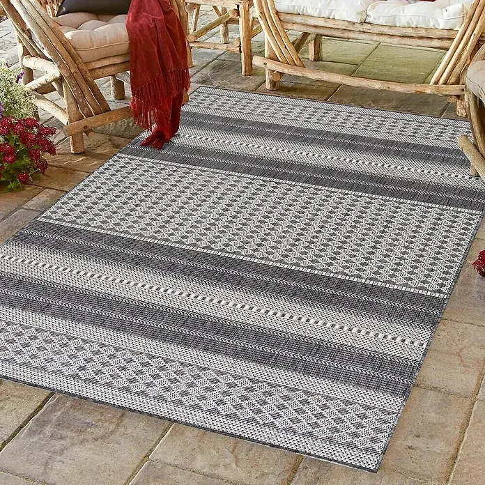 Outdoor-Teppich Sunny VI - Beige, 170 x 120 cm, 100 % Polypropylen - Ein rechteckiger grauer Teppich mit geometrischen Mustern liegt auf einer Terrasse mit Gartenmöbeln.