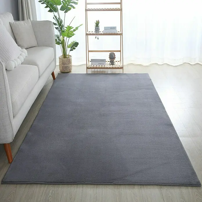 Kurzflorteppich Sky - Grau, 290 x 200 cm, 100 % Polyester - Grauer Teppich im Wohnzimmer. Der Teppich liegt vor einem hellen Sofa und einem Regal.