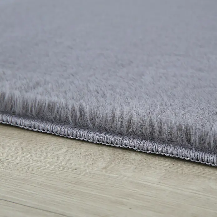 Hochflorteppich Pouffy - Grau, 290 x 200 cm, 100 % Polyester - Detailaufnahme eines flauschigen, grauen Teppichs mit einem ordentlichen, genähten Rand, der auf einem hellen Holzboden liegt.