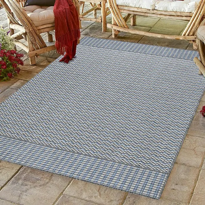 Outdoor-Teppich Sunny VII - Grau, 170 x 120 cm, 100 % Polypropylen - Ein blauer und grauer Outdoor-Teppich mit Bordüre liegt auf einer Steinterrasse unter Möbeln.