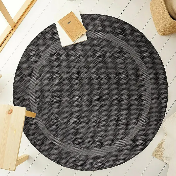 Outdoor-Teppich Relax - Schwarz, Durchmesser: 200 cm, 100 % Polypropylen - Ein runder, anthrazitfarbener Teppich mit einem hellgrauen Rand liegt auf einem weißen Holzboden und verleiht dem Raum ein gemütliches Flair.