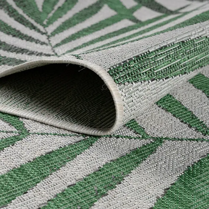 Outdoor-Teppich Sunny II - Grün, 170 x 120 cm, 100 % Polypropylen - Nahaufnahme eines modernen Teppichs mit grün-weißem Palmblattmuster, der teilweise aufgerollt ist und seine Textur zeigt.