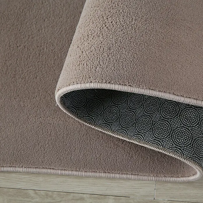 Kurzflorteppich Sky - Beige, 340 x 240 cm, 100 % Polyester - Detailaufnahme eines beigen Teppichs mit rutschfester Unterseite, der auf einem Holzboden liegt. Die Textur und Farbe des Teppichs sind gut erkennbar.