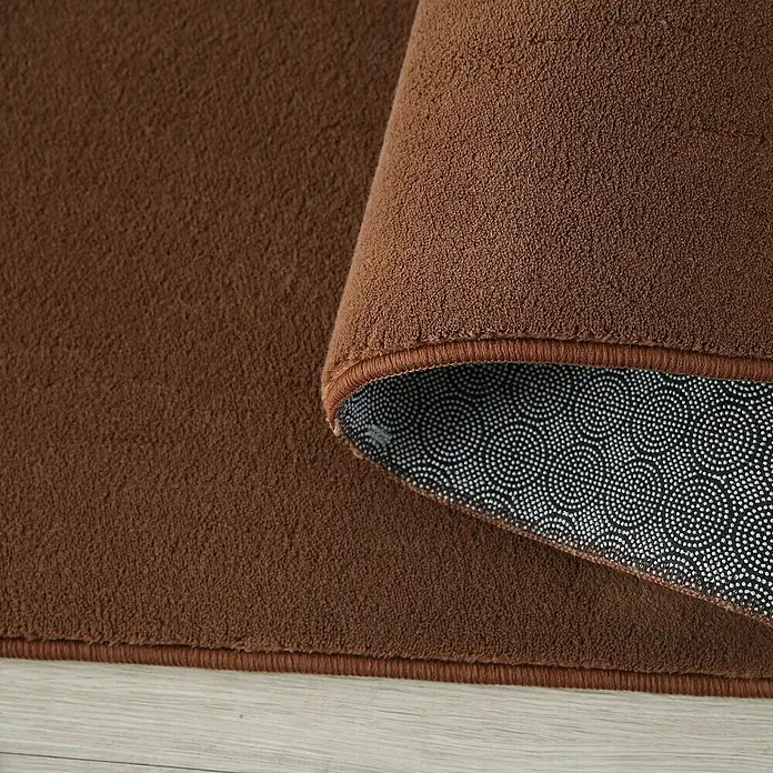Kurzflorteppich Sky - Braun, 200 x 140 cm, 100 % Polyester - Nahaufnahme eines braunen Teppichs mit gewellter Ecke, der die rutschfeste Unterseite zeigt.