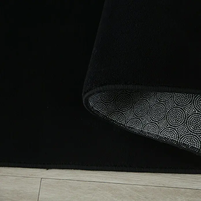 Kurzflorteppich Sky - Schwarz, 230 x 160 cm, 100 % Polyester - Nahaufnahme eines schwarzen Teppichs, der auf einem hellen Holzboden liegt und eine rutschfeste Unterseite mit Punktmuster zeigt.
