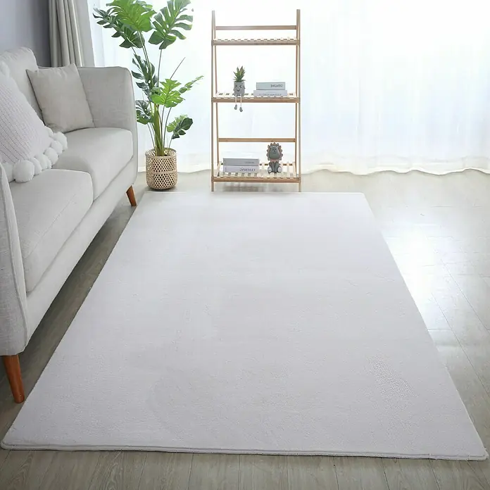 Kurzflorteppich Sky - Creme, 150 x 80 cm, 100 % Polyester - Ein weicher, weißer Teppich liegt in einem hellen Raum mit einem grauen Sofa, einem Regal und einer Zimmerpflanze.