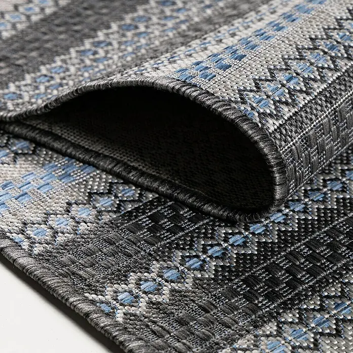 Outdoor-Teppich Sunny V - Schwarz, 170 x 120 cm, 100 % Polypropylen - Nahaufnahme eines gemusterten grauen Teppichs mit blauen Akzenten, der teilweise aufgerollt ist und seine Textur zeigt.