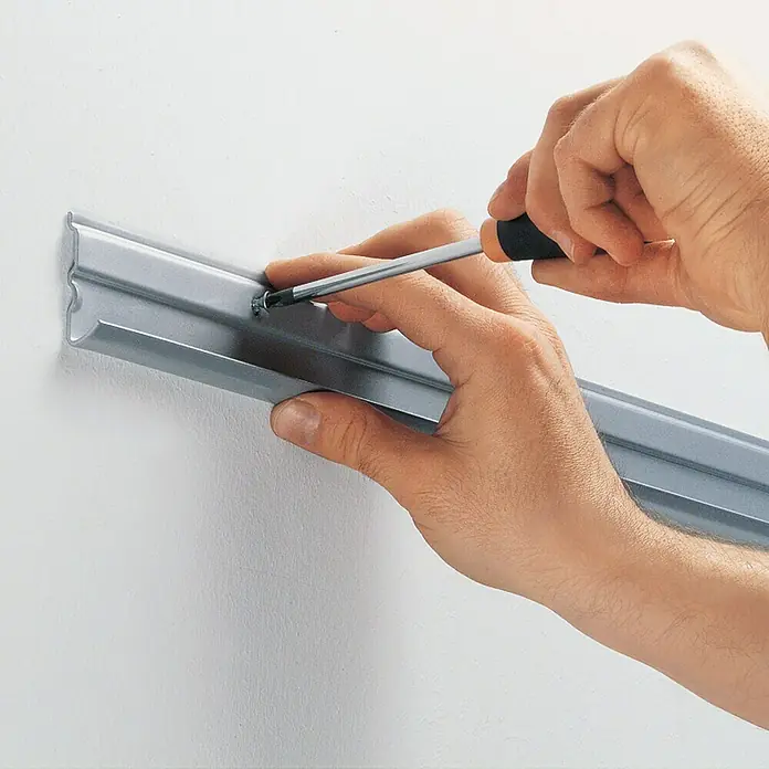 Element System Easy Hängeschiene 32 - Länge: 120 cm, Zweilochsystem - Nahaufnahme einer Person, die eine silberne Metallschiene mit einem Schraubenzieher an einer weißen Wand befestigt.