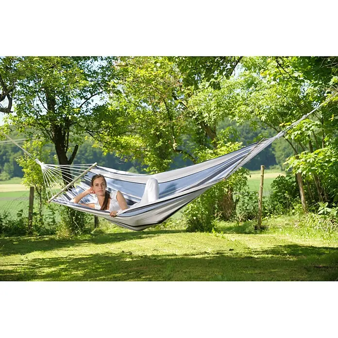 Amazonas Stabhängematte Samba marine - L x B: 210 x 140 cm, Blau/Weiß - Frau entspannt in einer gestreiften Hängematte im Garten, umgeben von Bäumen und Grünflächen. Die Hängematte bietet Komfort und Stil für den Außenbereich.