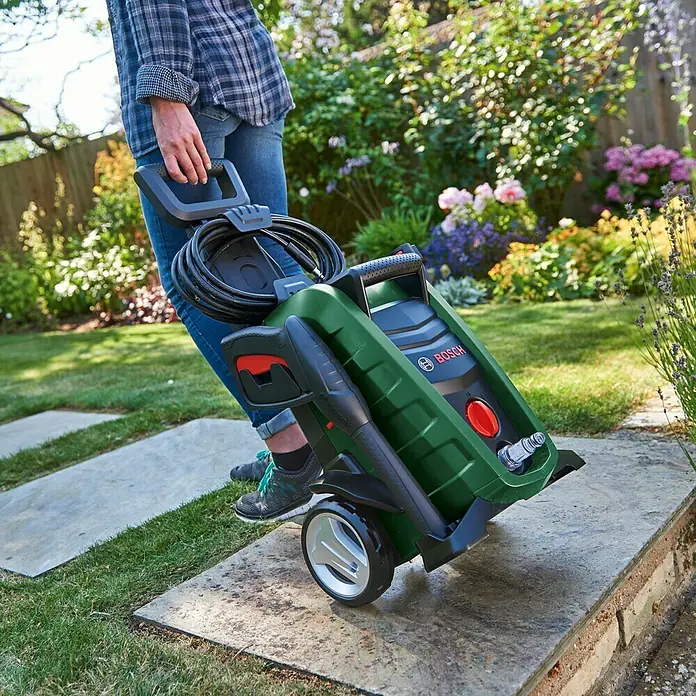 Bosch Hochdruckreiniger UniversalAquatak 130 - 1.700 W, Fördermenge: 380 l/h - Person, die einen grünen Bosch Hochdruckreiniger mit Rädern und einem Griff auf einer Terrasse zieht.