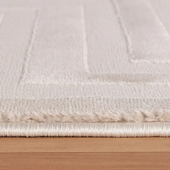 Kurzflorteppich Sahara V - Beige, 170 x 120 cm, 100 % Polypropylen - Detailaufnahme eines weißen Teppichs mit geometrischem Muster und weicher Textur, der auf einem Holzboden liegt.