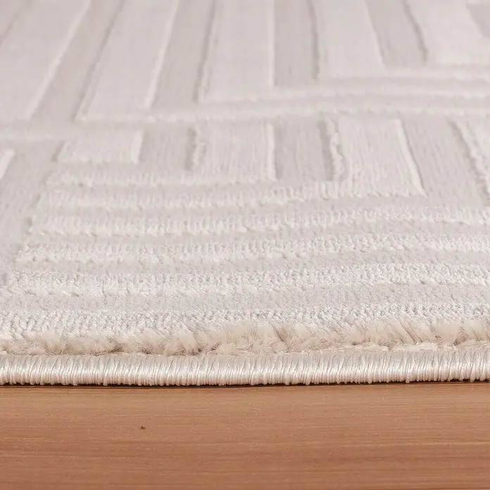Kurzflorteppich Sahara III - Beige, 170 x 120 cm, 100 % Polypropylen - Nahaufnahme eines weißen Teppichs mit geometrischem Muster auf einem Holzboden, der die Textur und das Design des Teppichs hervorhebt.