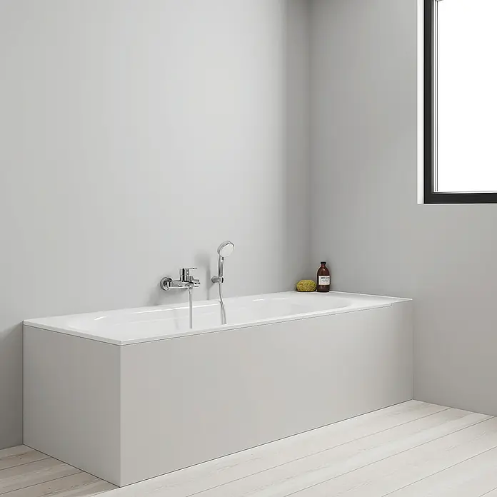 Grohe Eurostyle Cosmopolitan Badewannenarmatur - Ausladung Armatur: 173 mm, Chrom, Glänzend, Handbrause - Moderne Badewanne mit Armatur und Brause vor einer grauen Wand und einem Fenster. Der Boden ist helles Holz.