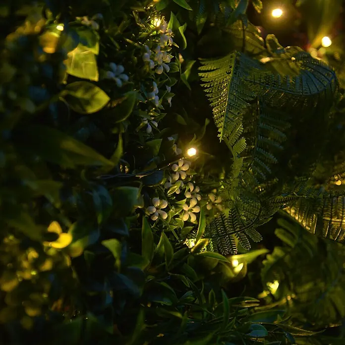 Jardín vertical Jungle con luces LED - Plástico, L x An: 100 x 100 cm - Primer plano de un exuberante panel de pared de plantas artificiales con pequeñas flores blancas y luces cálidas que crean un ambiente acogedor.