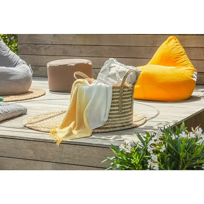 Juteteppich Nature 502 - Naturbraun, Durchmesser: 100 cm, 100 % Jute - Ein dekorativer Korb mit einem Kissen und einer Decke auf einer hölzernen Terrasse, umgeben von Sitzsäcken und Blumen.