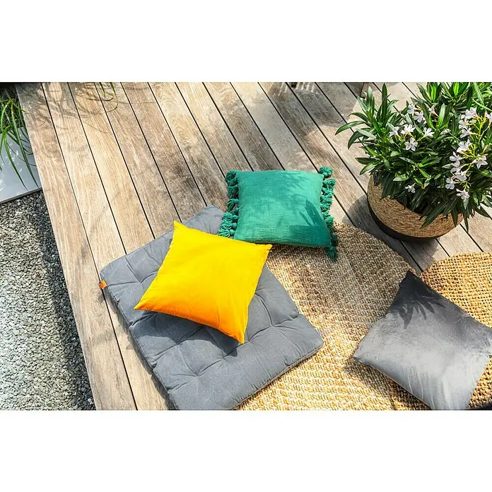 Juteteppich Nature 502 - Naturbraun, Durchmesser: 100 cm, 100 % Jute - Außenbereichsszene mit dekorativen Kissen in Gelb, Grün und Grau auf einem runden Teppich, neben einem Topf mit grünen Pflanzen und weißen Blüten.