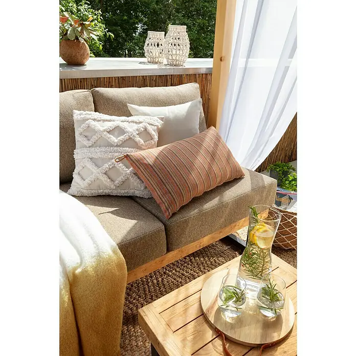 SENSUM Kissen - Bunt, L x B: 65 x 30 cm - Nahaufnahme eines entspannenden Außenbereichs mit beigen Kissen, einem gestreiften Dekokissen und einem Getränkeset mit Zitronen auf einem Holztisch.