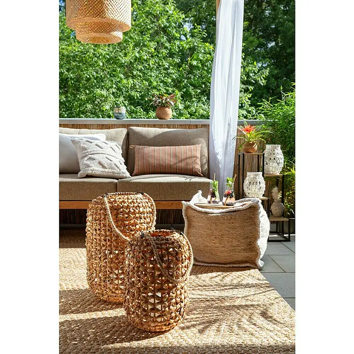 SENSUM Kissen - Bunt, L x B: 65 x 30 cm - Gemütlicher Balkonbereich mit zwei dekorativen Laternen aus Naturfasern, einem Pouf und einem Outdoor-Sofa vor üppigem Grün.
