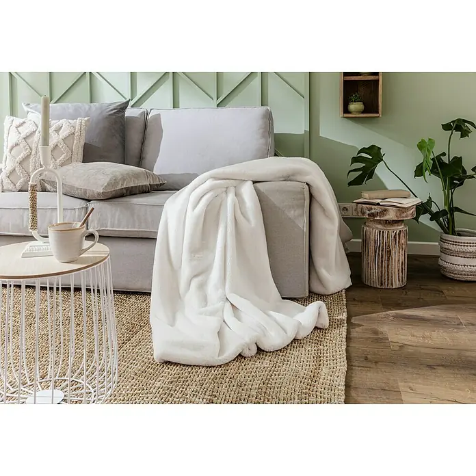 Decke HappyElfenbein, 200 x 150 cm, 100 % Polyester - Nahaufnahme einer weichen, weißen Decke, die elegant über eine graue Couch drapiert ist, neben einem Beistelltisch und einer Pflanze.