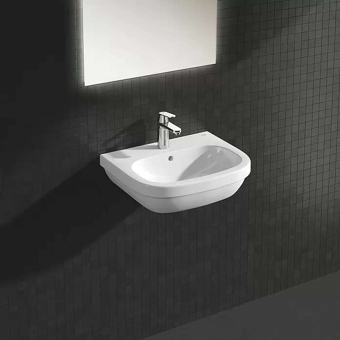 Grohe Eurosmart Cosmopolitan Waschtischarmatur M-Size - Mit Zugstange, Chrom, Glänzend - Weißes wandmontiertes Waschbecken mit Chromarmatur, vor einer dunklen Fliesenwand unter einem beleuchteten Spiegel.