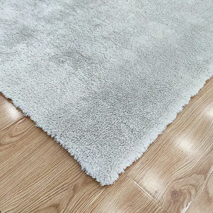 Alfombra de pelo largo Bolonia - Blanco, 115 x 60 cm - Primer plano de una alfombra de baño suave de color gris claro sobre un suelo de madera, que combina confort y estilo.