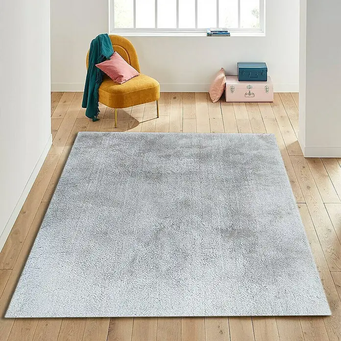 Alfombra de pelo largo Bolonia - Blanco, 115 x 60 cm - Una alfombra gris está tendida en un piso de madera en una habitación luminosa, creando un ambiente acogedor. La alfombra es el elemento central de la habitación.