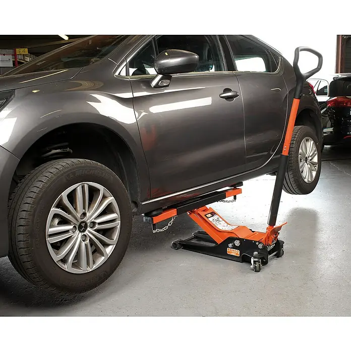 Bahco Querträger BH1CB1000Passend für: Bahco Wagenheber H1M1000/BH11500/BH12000/BH13000 - Nahaufnahme eines grauen Autos, das mit einem orangefarbenen Bahco-Rangierwagenheber für Reparaturarbeiten in einer Werkstatt angehoben wird.