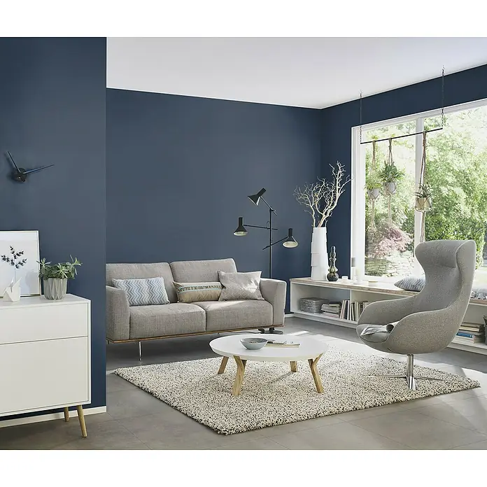 Rasch Tropical House Vliestapete - Nachtblau, Uni, 10,05 x 0,53 m - Ein stilvolles Wohnzimmer mit einem grauen Sofa, einem Sessel, einem weißen Couchtisch und einem weißen Sideboard. Die Wände sind in einem tiefen Blau gehalten.