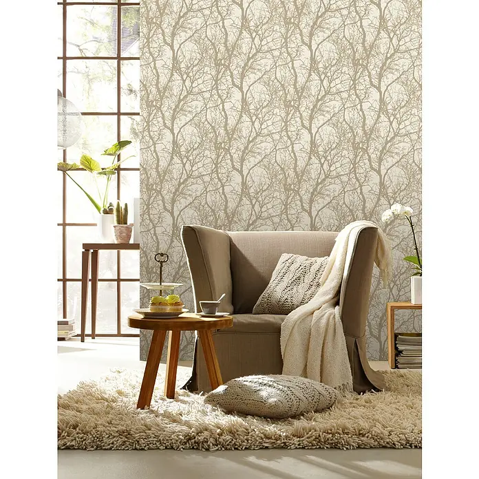Rasch Tapetenwechsel Vliestapete Äste - Beige/Bronze, Floral, 10,05 x 0,53 m - Beige Sessel mit Kissen und Decke, platziert auf einem beigen Teppich vor einer Wand mit abstraktem Baumdesign. Einladende und gemütliche Atmosphäre.