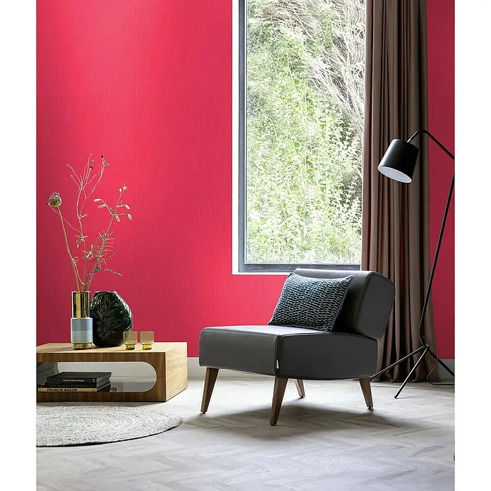 Rasch Wall Textures VI Vliestapete Struktur-Uni - Dunkelpink, Uni, 10,05 x 0,53 m - Elegantes Interieur mit einem modernen grauen Sessel, einem stilvollen Holztisch und einer hohen Stehlampe vor einer roten Wand.