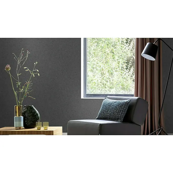 Rasch Factory V Vliestapete Beton-Uni - Offblack, Uni, 10,05 x 0,53 m - Gemütlicher Wohnraum mit dunklen Akzenten, einem grauen Sessel mit Kissen und einer Stehlampe neben einem Fenster mit Vorhängen.