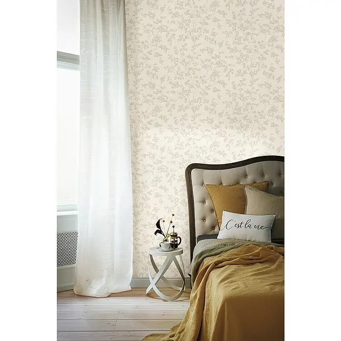 Rasch Welcome home Vliestapete Pflanzen - Greige, Floral, 10,05 x 0,53 m - Schlafzimmerdekor mit einer beigen, blattgemusterten Tapete, ergänzt durch ein stilvolles Bett und einen eleganten Beistelltisch.