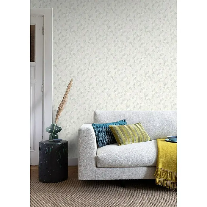 Rasch Welcome home Vliestapete Grafisch - Hellgrau, Grafisch, 10,05 x 0,53 m - Das Bild zeigt ein modernes Wohnzimmer mit einem stilvollen Sofa, Kissen und einer dekorativen Vase auf einem Beistelltisch. Die Wand ist mit einer Mustertapete verziert.