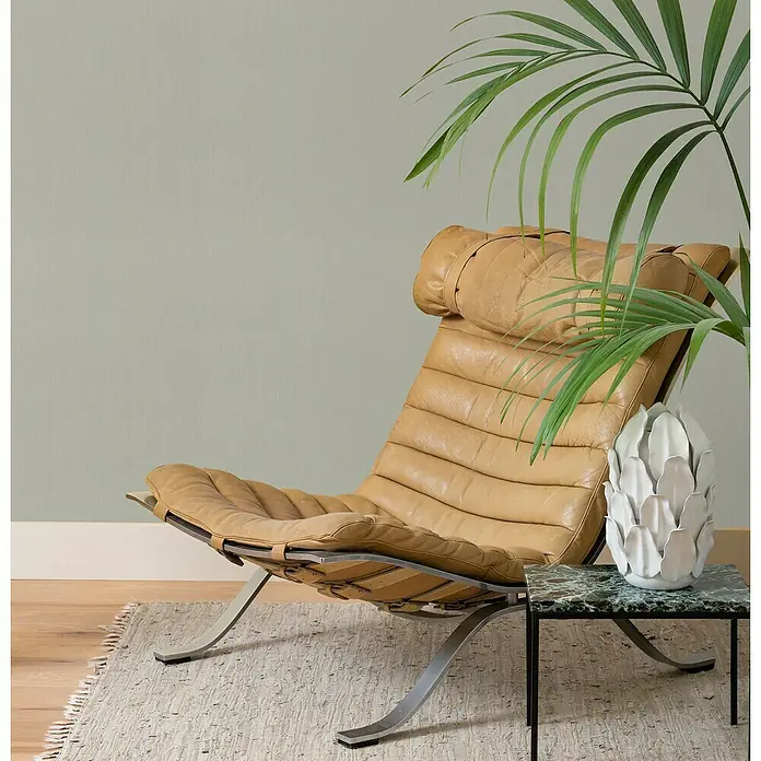 Rasch Wall Textures VI Vliestapete Metallic-Uni - Samtgrau, Uni, 10,05 x 0,53 m - Elegante Ledersessel mit Stahlrahmen und ein Marmortischchen mit dekorativer Vase, umgeben von einer tropischen Pflanze, die ein entspanntes Ambiente erzeugen.