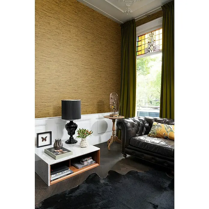 Rasch Curiosity Vliestapete Holzoptik - Gold, Holzoptik, 10,05 x 0,53 m - Elegant eingerichtetes Wohnzimmer mit dunklem Ledersofa und stilvoller Dekoration. Die goldfarbene Tapete und die grünen Vorhänge setzen Akzente.