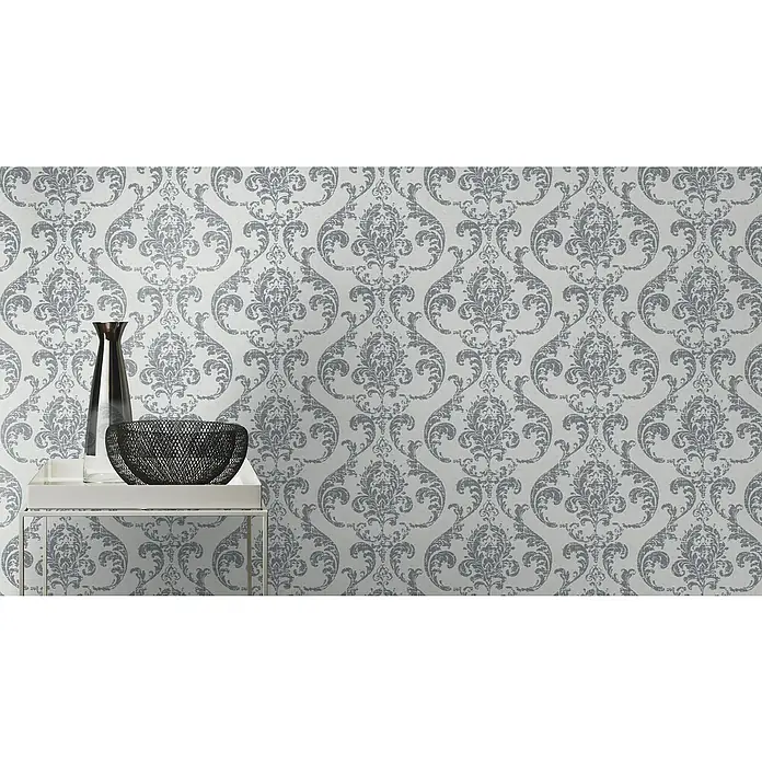 Rasch Color your life Vliestapete Ornamente - Hellgrau, Grafisch, 10,05 x 0,53 m - Einrichtungsfoto mit einer schlanken Vase und einer dekorativen Schale auf einem weißen Tisch vor einer Wand mit grauem Damastmuster.