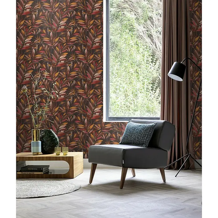 Rasch Symphony Vliestapete Blätter - Dunkelrot, Floral, 10,05 x 0,53 m - Ein grauer Sessel mit Kissen vor einem Fenster mit Vorhängen. Eine Lampe steht daneben, und ein Tisch mit Dekorationen vervollständigt das Bild.