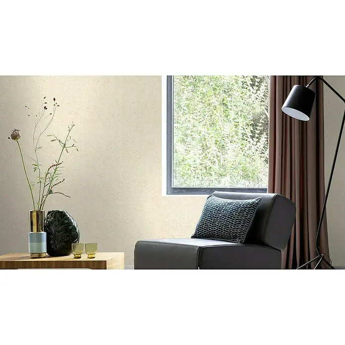 Rasch Factory V Vliestapete Beton-Uni - Greige, Uni, 10,05 x 0,53 m - Das Bild zeigt eine moderne Wohnzene mit einem stilvollen Sessel, dekoriert mit einem blauen Kissen, neben einem Fenster und einer Stehlampe.