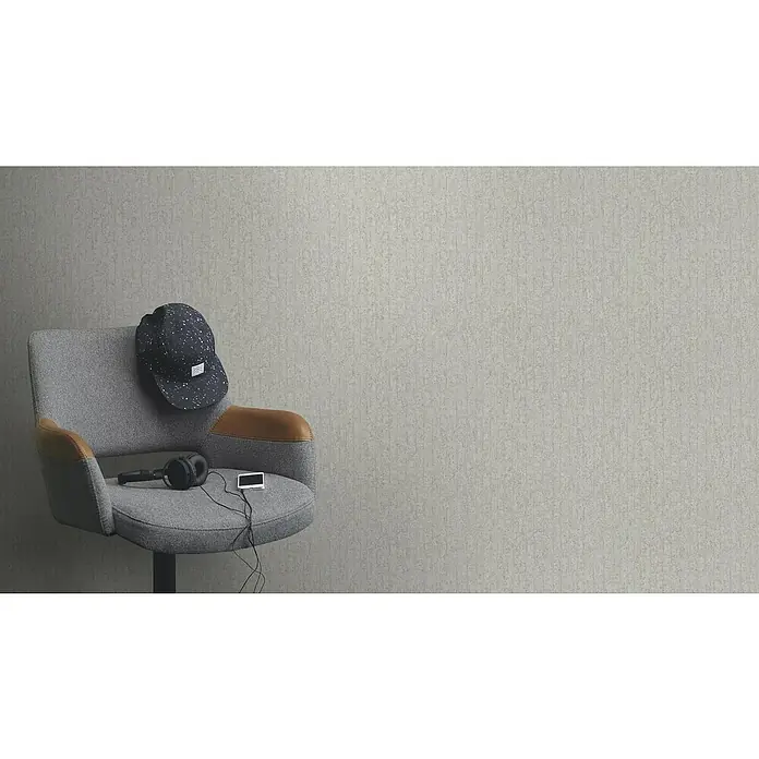 Rasch Factory V Vliestapete Betonoptik-Uni - Greige, Steinoptik, 10,05 x 0,53 m - Nahaufnahme eines grauen Drehstuhls mit Kopfhörern, Kappe und einem Gerät. Der Stuhl hat Lederakzente und steht vor einer grauen Wand.