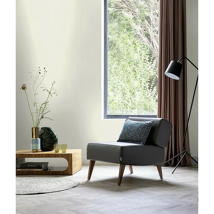 Rasch Sky Lounge Vliestapete Metallic-Uni - Perlmutt, Uni, 10,05 x 0,53 m - Ein moderner, grauer Sessel mit Holztisch und Dekorationen vor einem Fenster mit Vorhängen.