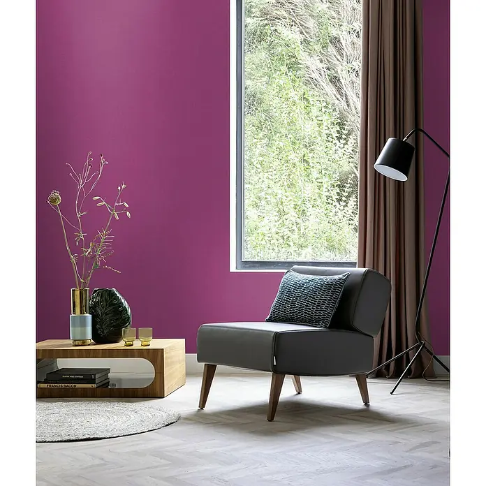 Rasch Country Charme Vliestapete Textil-Uni - Pink, Uni, 10,05 x 0,53 m - Ein stilvoller grauer Sessel mit blauem Kissen vor einer violetten Wand, ergänzt durch einen Holztisch und eine Stehlampe, schafft eine moderne Atmosphäre.