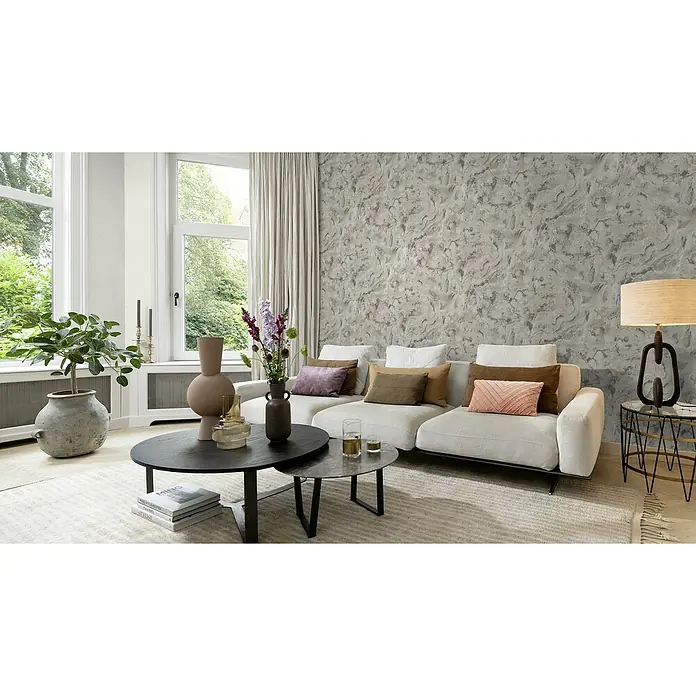 Rasch Symphony Vliestapete Marmor - Mittelgrau, Steinoptik, 10,05 x 0,53 m - Einrichtung eines modernen Wohnzimmers mit einem modularen Sofa, das mit Zierkissen und zwei Couchtischen dekoriert ist.