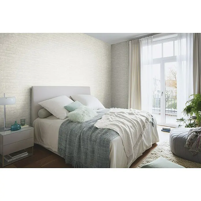 Rasch Factory V Vliestapete Beton - Greige, Steinoptik, 10,05 x 0,53 m - Schlafzimmer mit weiß getünchter Ziegelwand, Bett mit weichen Kissen und Tagesdecke, Nachttisch mit Lampe und Fenster mit Vorhängen.