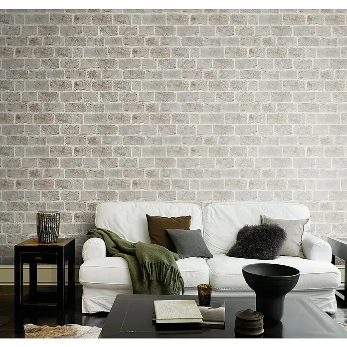 Rasch Symphony Vliestapete Backstein - Steingrau, Steinoptik, 10,05 x 0,53 m - Gemütliches Wohnzimmer mit einer weißen Couch, verschiedenen Kissen und einer stilvollen Dekoration vor einer Backsteinwand.