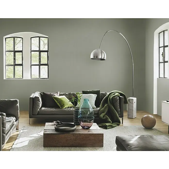 Rasch African Queen III Vliestapete Lederoptik - Olivgrün, Uni, 10,05 x 0,53 m - Elegantes Wohnzimmer mit einem grauen Ledersofa, einer Stehlampe und einem Couchtisch aus Holz. Die Dekoration ist stilvoll und modern.