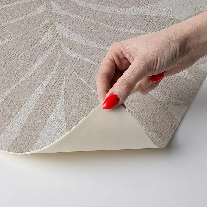 Alfombra de vinilo Tropikal - Crema, 110 x 48 cm, PVC - Primer plano de un papel pintado autoadhesivo con estampado de hojas que está siendo retirado por una mano con esmalte de uñas rojo.