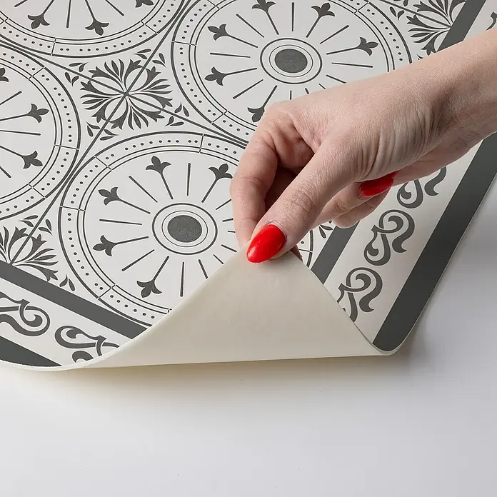 Alfombra de vinilo Buzz - Gris, 75 x 48 cm, PVC - Primer plano de una mano levantando un pedazo de suelo de vinilo con un diseño de patrón gris y blanco.